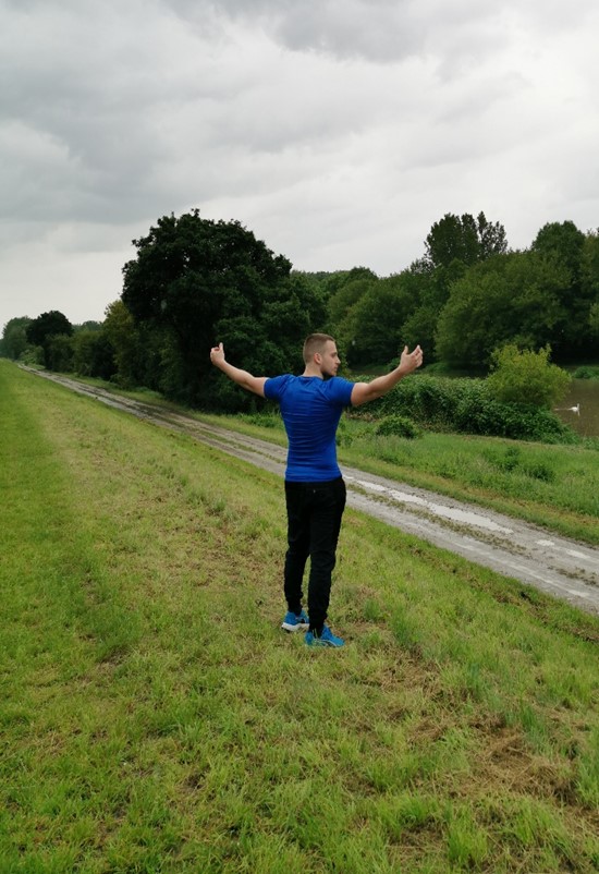 Der Eigentümer der Website, Milos, befindet sich in einer Jogginghose mit einem Trainingsshirt auf einem Grasweg nahe eines schmalen Flusses in der Nähe von Wien. Er streckt seine Arme auseinander und schaut in Richtung des Flusses. Im Hintergrund ist der Fluss von Bäumen und Sträuchern umgeben. Der Himmel ist stark bewölkt, aber dennoch hell.